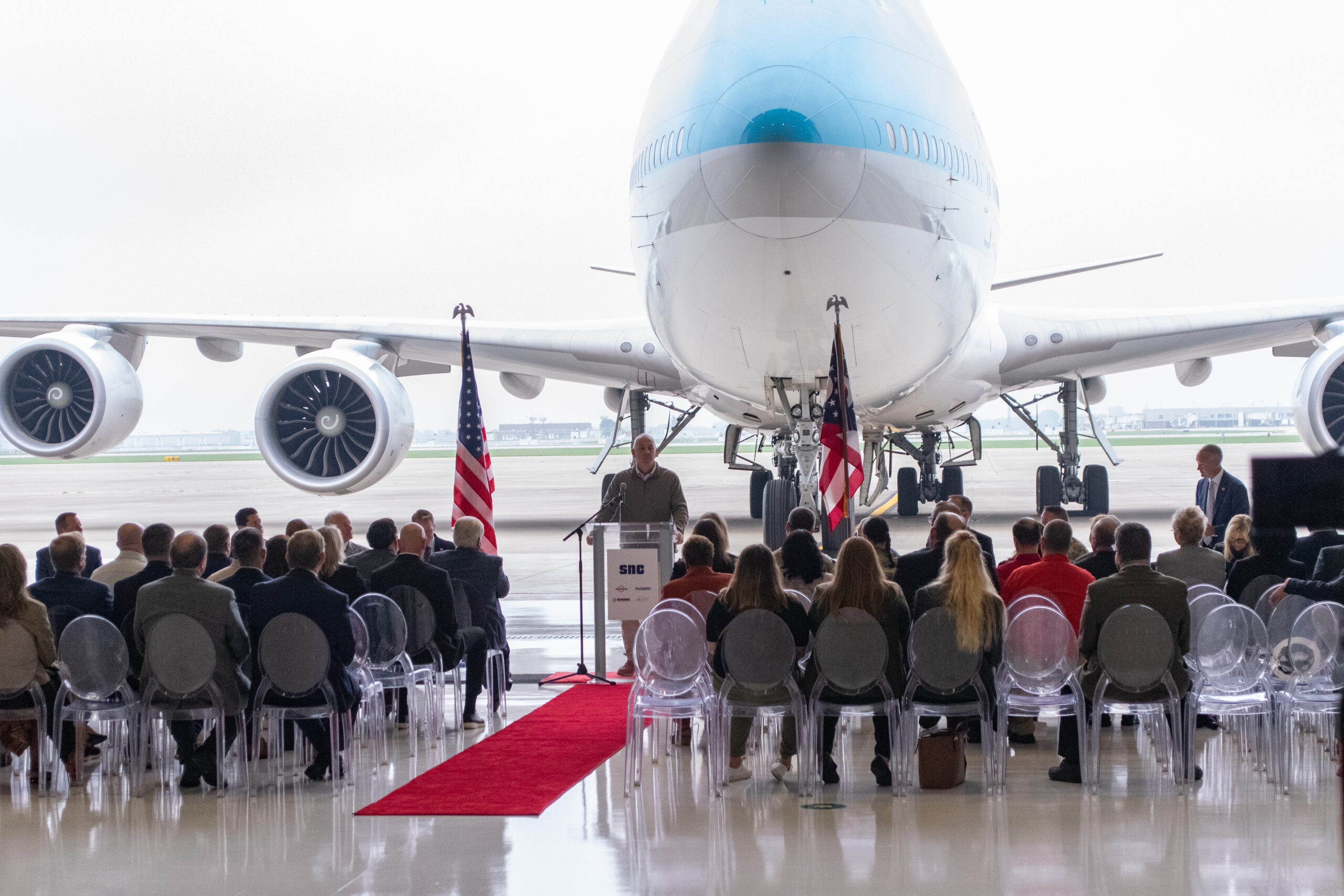 A speaker addresses an audience seated in front of a large airplane in a hangar, with American flags and a red carpet leading to the podium.