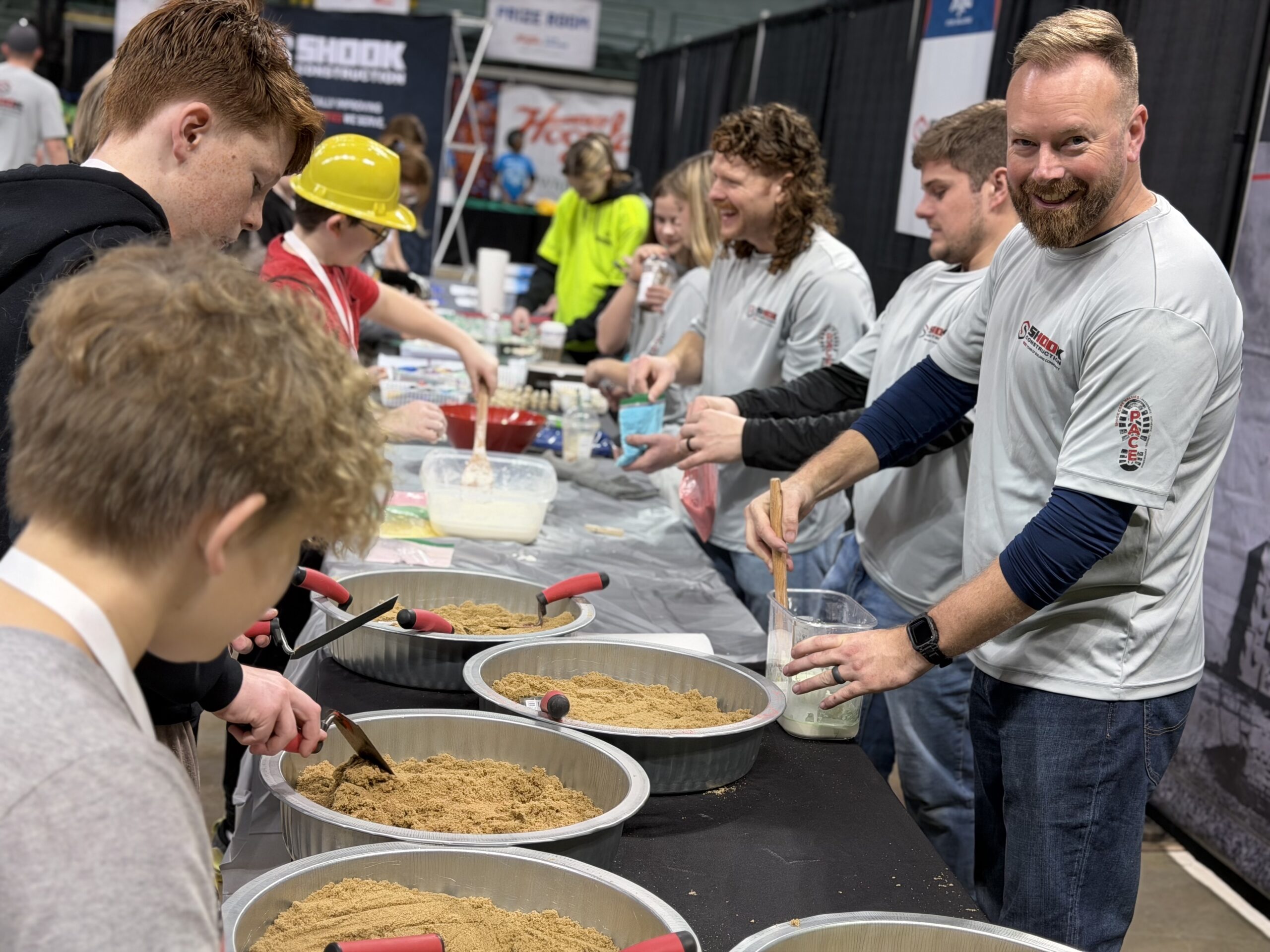 Adults and kids gather around tables filled with sand, working on hands-on projects at an indoor event. Adults assist children, while others in the background observe and participate.