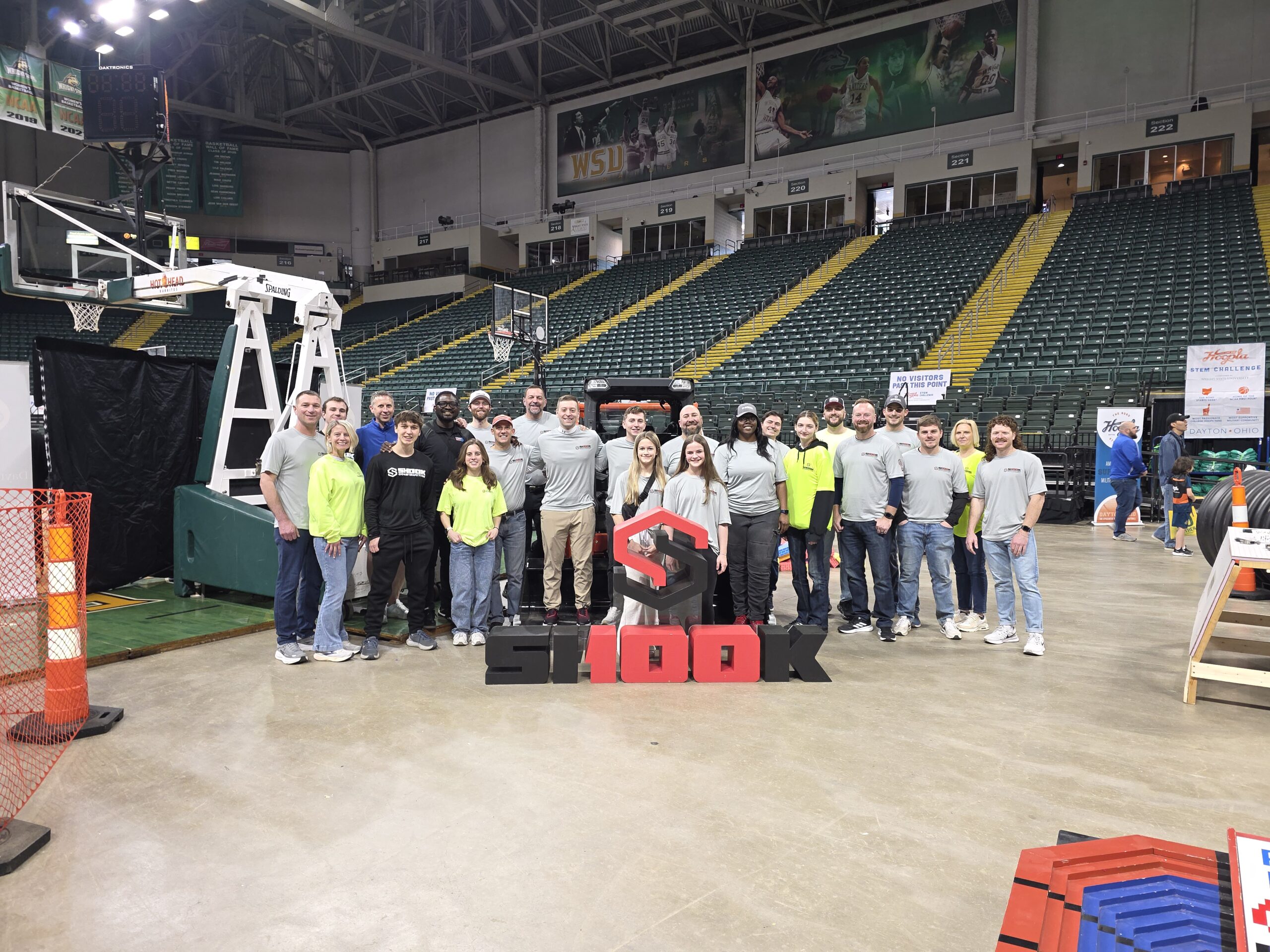 A large group of people pose on a gym floor, some in gray shirts and others in neon yellow. They stand behind a red and black SHOOK sign, with empty green stadium seats and a basketball hoop in the background.