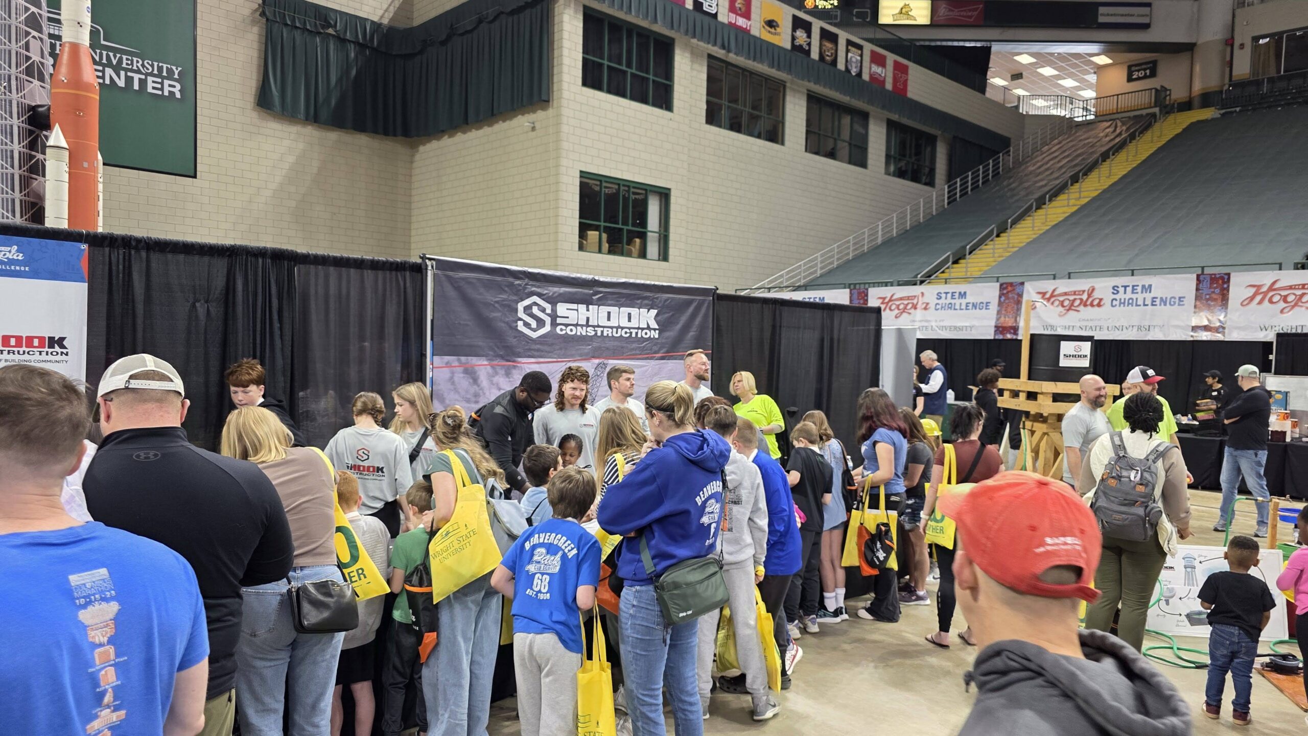 A crowd of people, many wearing blue shirts and holding yellow bags, gathers at a STEM event in a large indoor arena near a booth with a Shook Construction banner.
