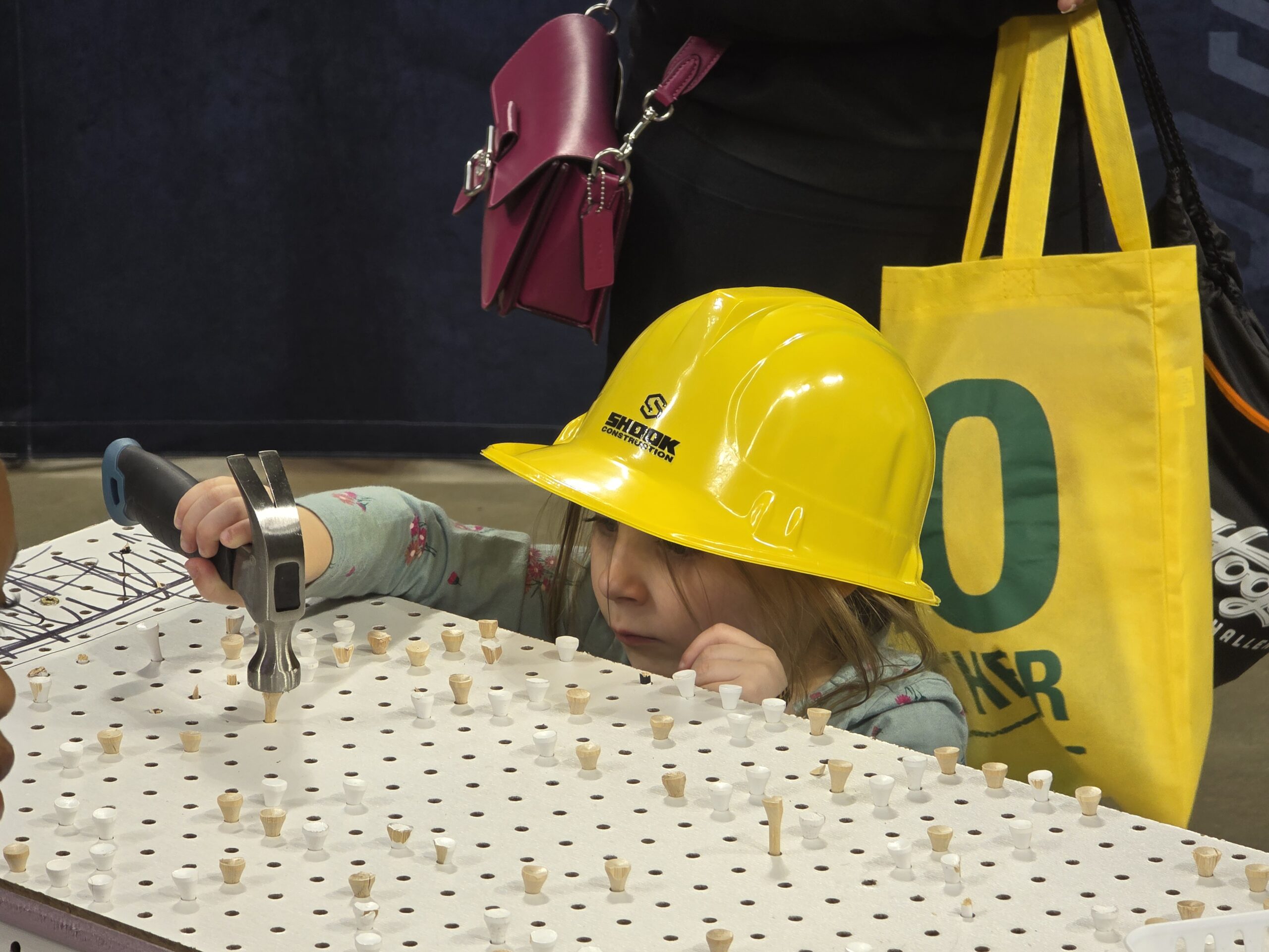 A young child wearing a yellow hard hat uses a hammer on a pegboard. Adults stand nearby, one holding a bright yellow tote bag and another with a pink purse.