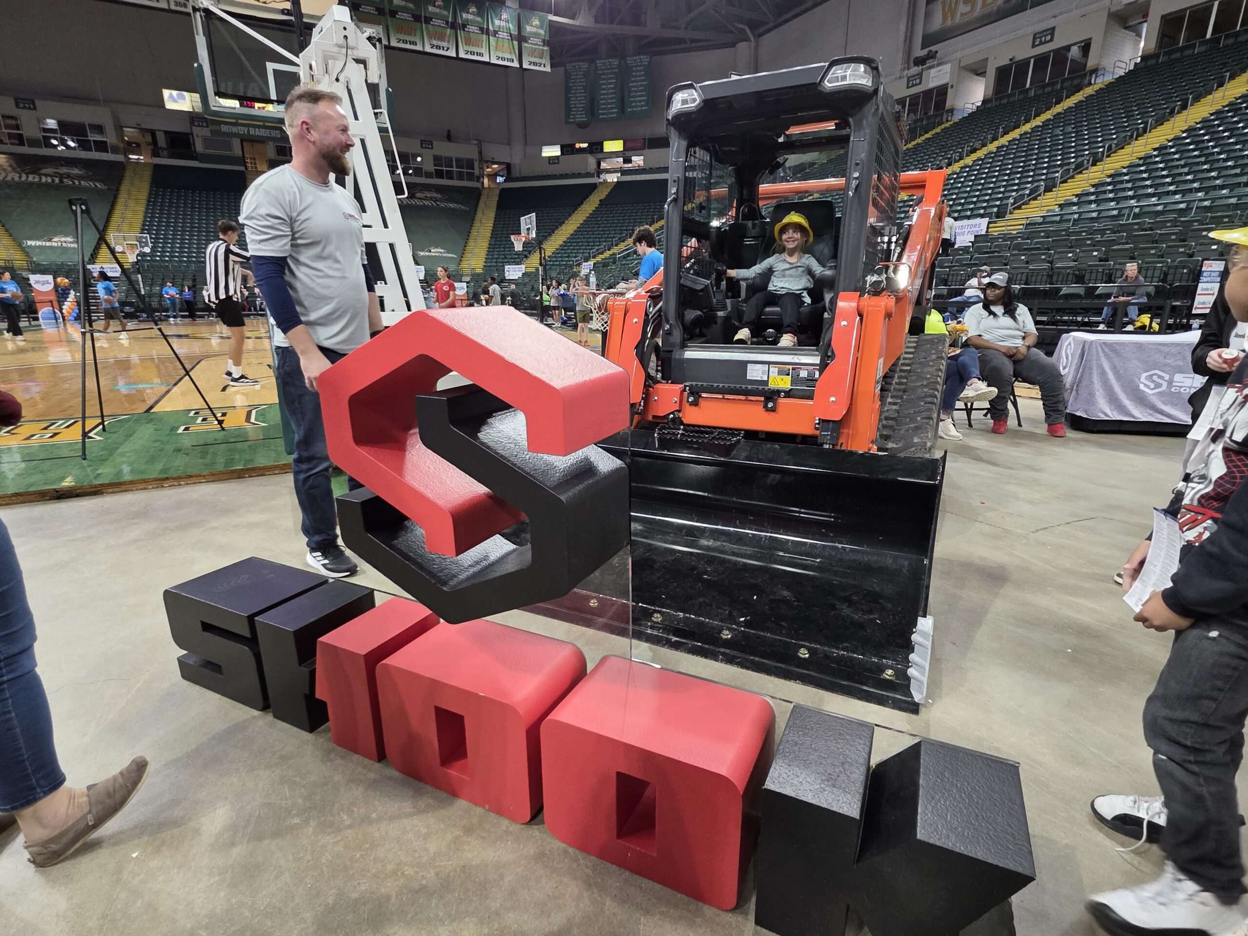 A child in a construction vehicle smiles while an adult stands nearby. Large block letters in front spell SIOUX. The scene is inside an arena with people and equipment in the background.