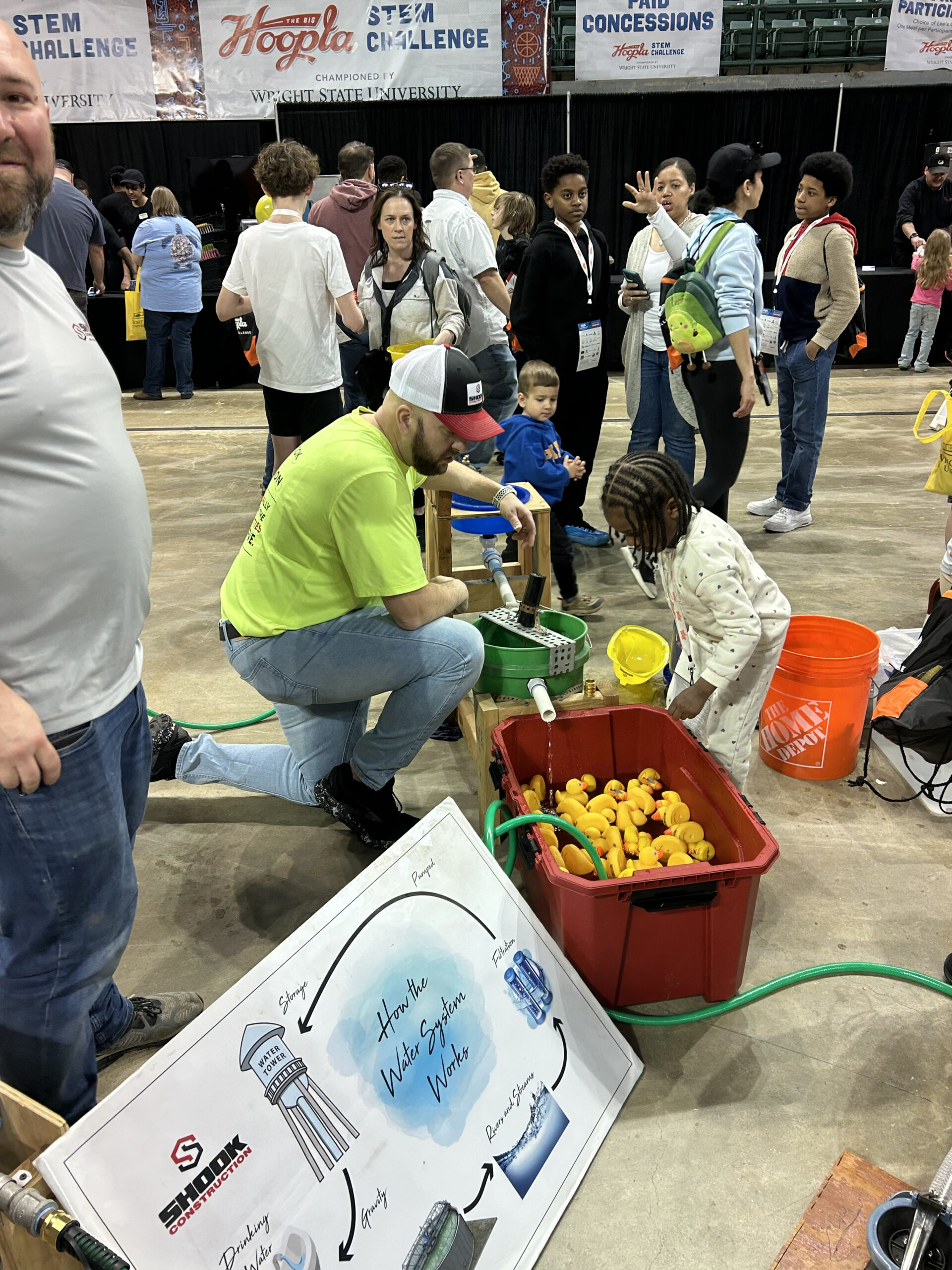 A man in a neon shirt kneels by a tub with toy ducks, showing a young girl how to use a water pump. People and children are gathered around in a busy indoor STEM event. A science diagram is visible in the foreground.