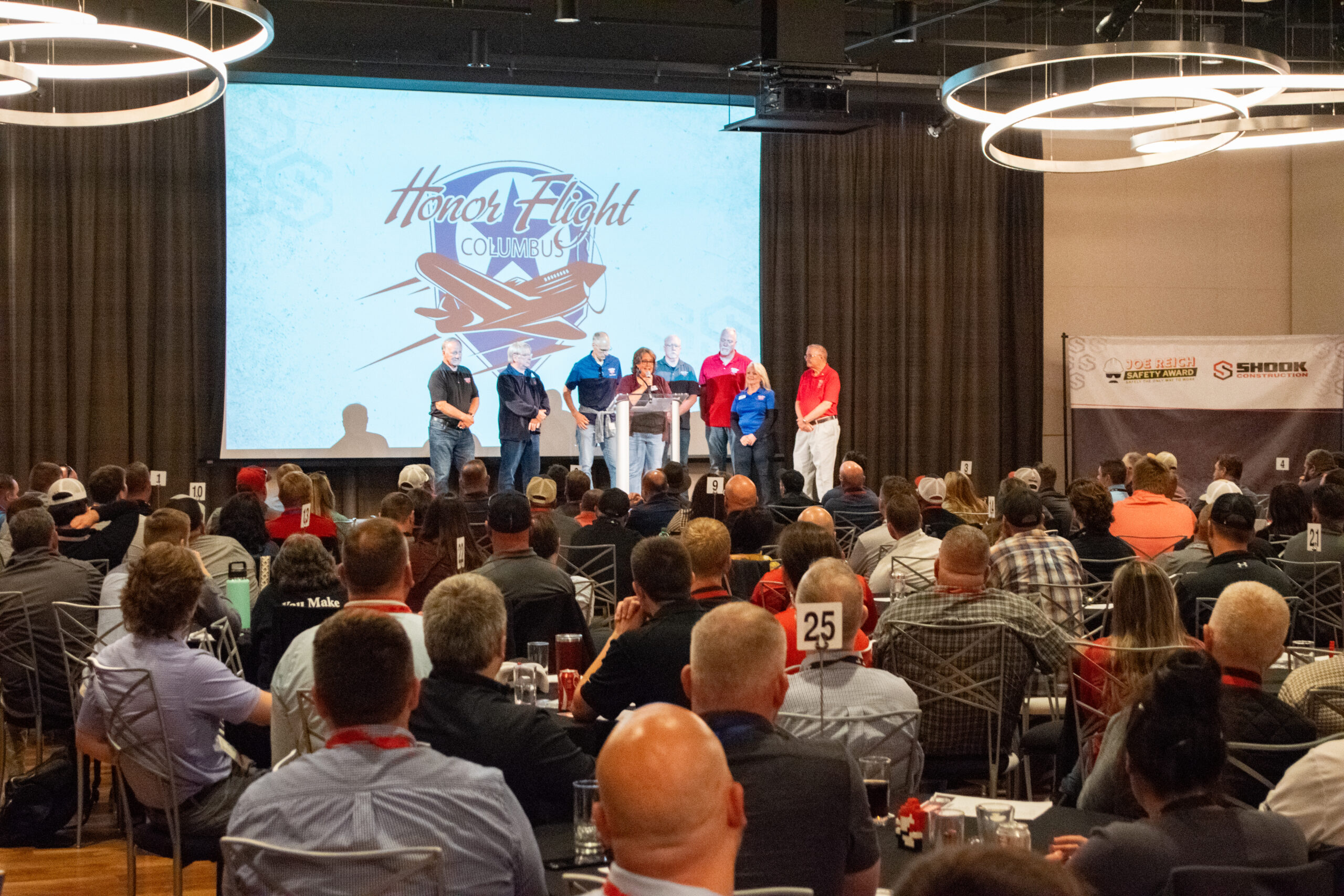 A group of people stand on stage speaking at an Honor Flight Columbus event while a large audience is seated, watching them in a conference room.