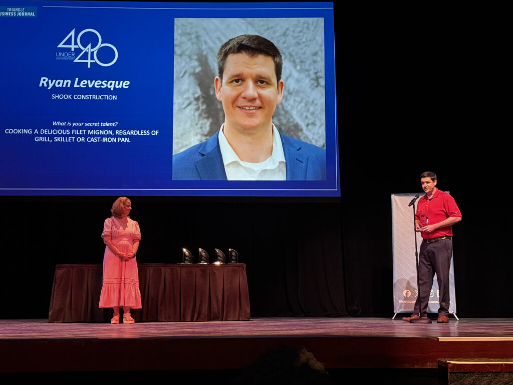 A man speaks at a podium on stage next to a woman. Behind them is a screen showing a photo of the man, his name (Ryan Levesque), and a 40 Under 40 award graphic.