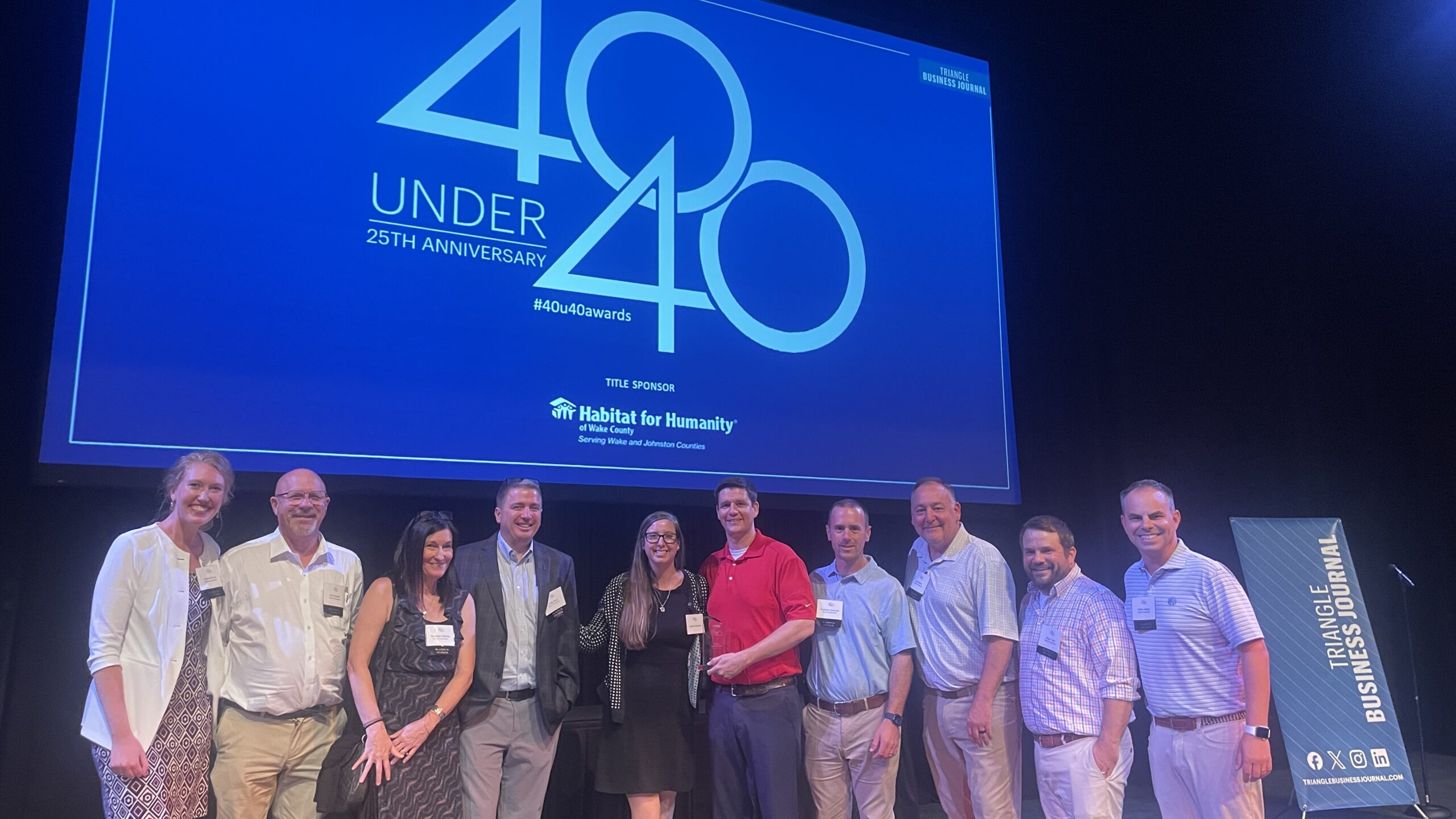 A group of eleven people poses on stage in front of a large 40 Under 40 25th Anniversary sign at a Habitat for Humanity event.