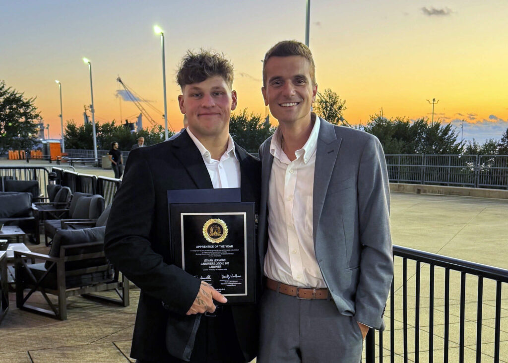 Two men in suits stand outside at sunset. One man holds a framed award plaque as they smile at the camera. Trees, outdoor seating, and lights are in the background under a colorful sky.