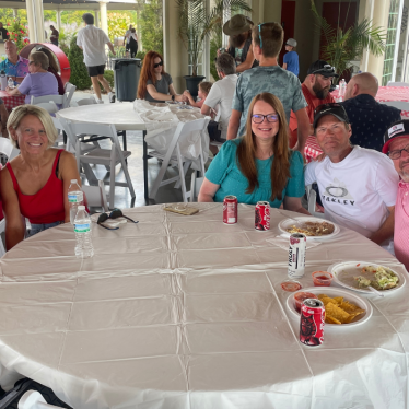 Five people sit smiling at a round table with food and drinks at a casual indoor event. Other guests and tables appear in the background, with sunlight streaming through large windows.