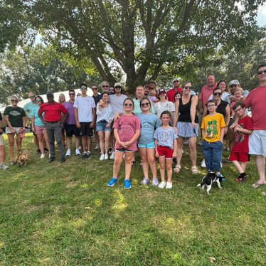 A large group of people, including children and adults, pose for a photo outdoors on grass under a tree. Some are wearing sunglasses, and two dogs are present. It is a sunny day.