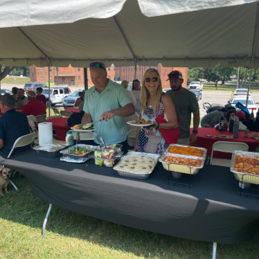 People serve themselves food from a buffet table under a large tent at an outdoor gathering. Others sit at tables in the background, and a small dog is visible near the buffet table.