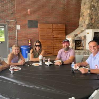 Five people sit around a table with food and drinks, smiling at the camera. Theres a brick wall, stacked wood, and a stone fireplace in the background, with sunlight coming in from a nearby open area.