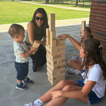 A woman and three children play with large wooden Jenga blocks outside on a patio. The girl in front places a block on top while the toddler holds a block. The group appears focused and happy.