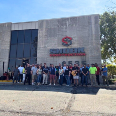 A large group of people stand in front of a building with a Shook Construction sign on the facade. Some people are holding musical instruments. It is a sunny day.