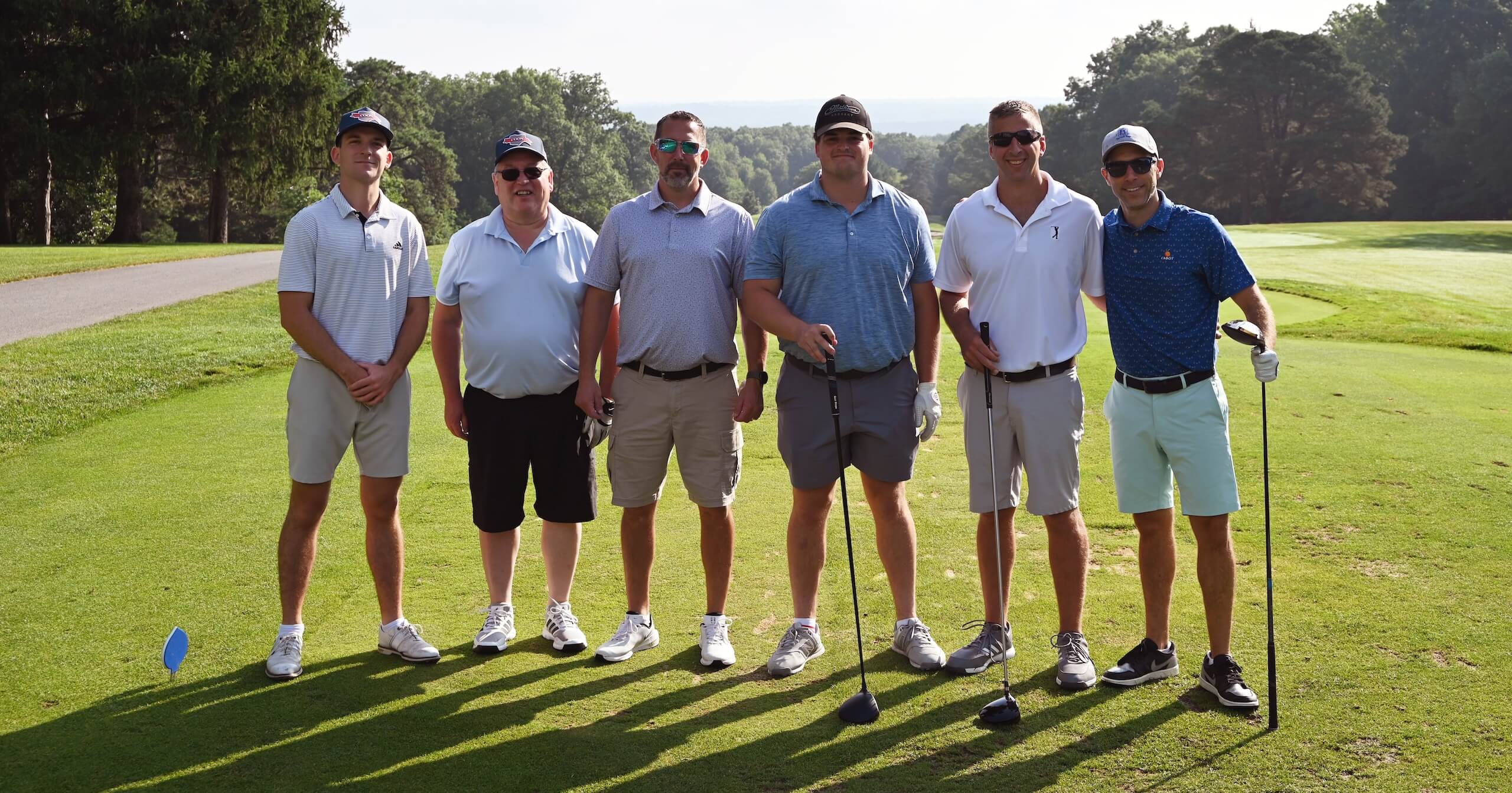 Seven men stand side by side on a golf course, each holding or near golf clubs, dressed in casual golf attire and hats, with trees and a sunny landscape in the background.