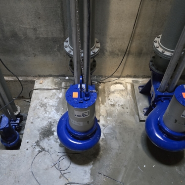 Three large blue and silver industrial pumps installed on a concrete floor, with vertical metal pipes attached, in a dimly lit utility or mechanical room. Loose wires and a chain are visible around the equipment.