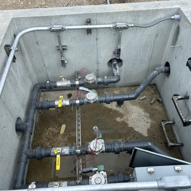 Concrete utility box containing multiple black pipes with valves, gauges, and labels; puddles of water on the floor; various cables and conduit attached to the walls; outdoor setting.