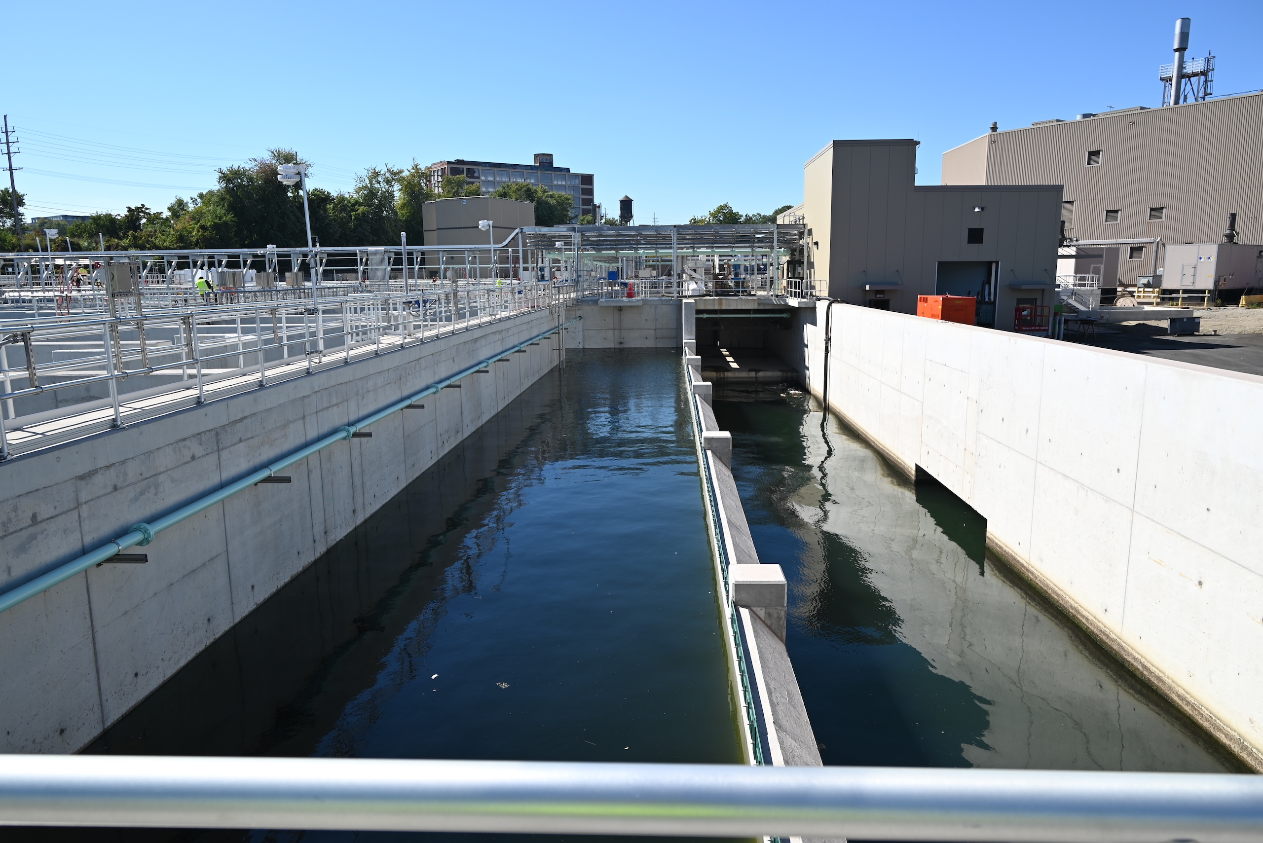 Outdoor view of a modern water treatment facility, featuring concrete channels filled with water, metal railings, and industrial buildings under a clear blue sky.