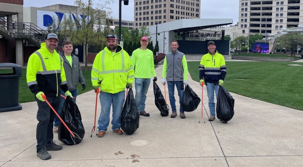 Six people stand on a sidewalk holding litter pickers and large trash bags, wearing work clothes and smiles, with buildings and grass in the background.