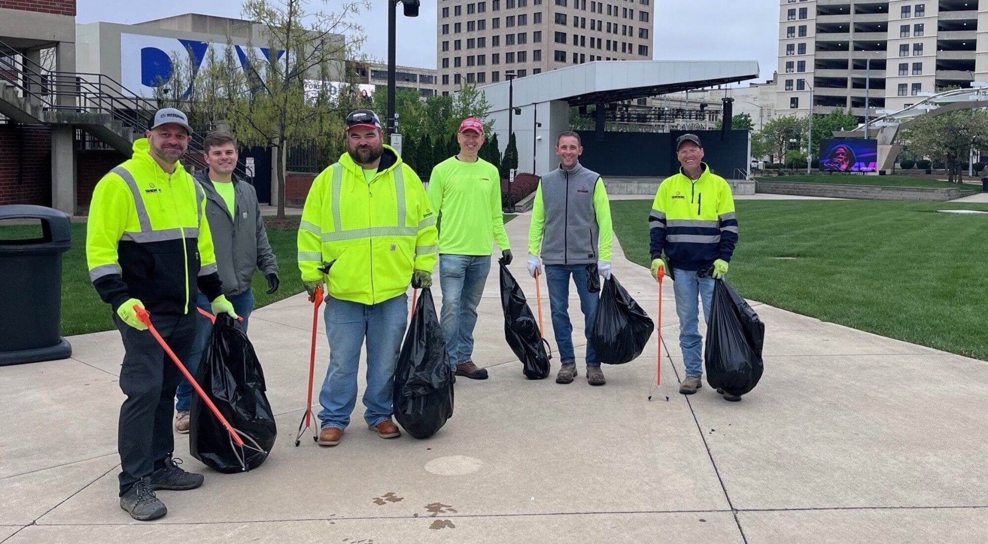 Six people stand on a sidewalk holding litter pickers and large trash bags, wearing work clothes and smiles, with buildings and grass in the background.