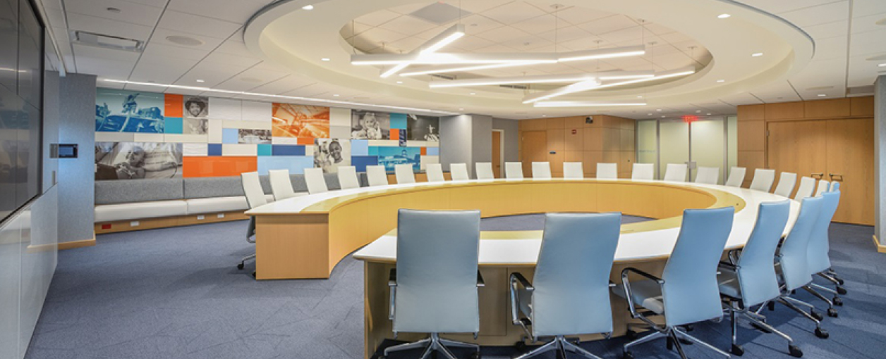 Modern conference room with a large oval table, white chairs, blue carpet, and a colorful mural on the back wall. Overhead lighting is sleek and contemporary. The space appears bright and organized for meetings.