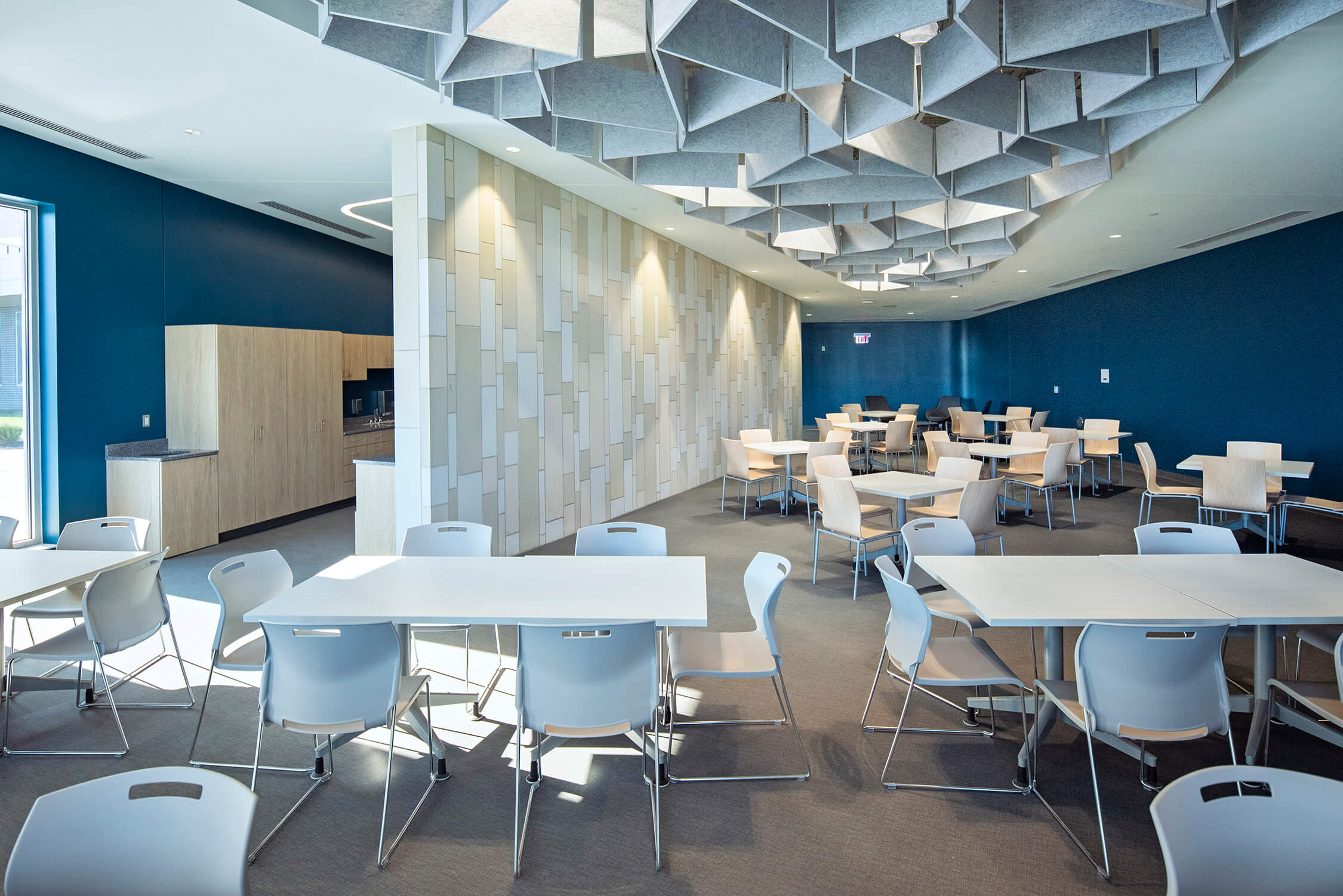 Modern cafeteria with light wooden tables and chairs, blue walls, and a geometric ceiling design. The room is bright, spacious, and minimally decorated, with large windows letting in natural light.