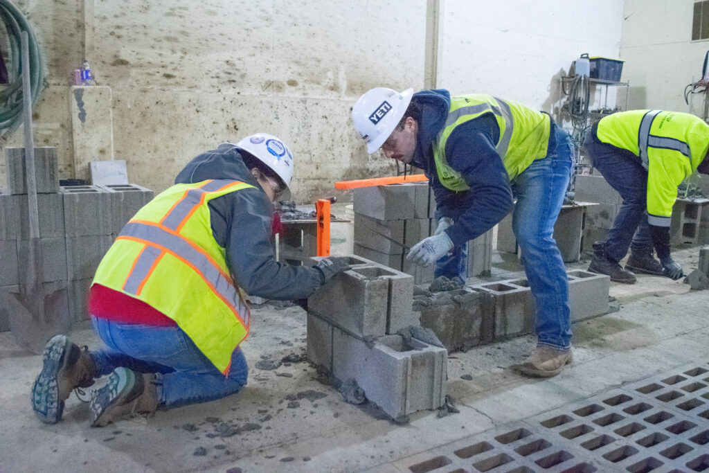 Two people in safety gear and hard hats lay concrete cinder blocks indoors, while another person in a yellow vest works nearby. The scene appears to be part of a construction or masonry activity.