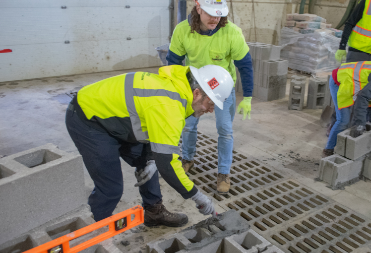 Two construction workers wearing safety gear and helmets lay concrete blocks on the floor inside a building. One uses a trowel, while the other observes. Bags of materials and tools are visible in the background.