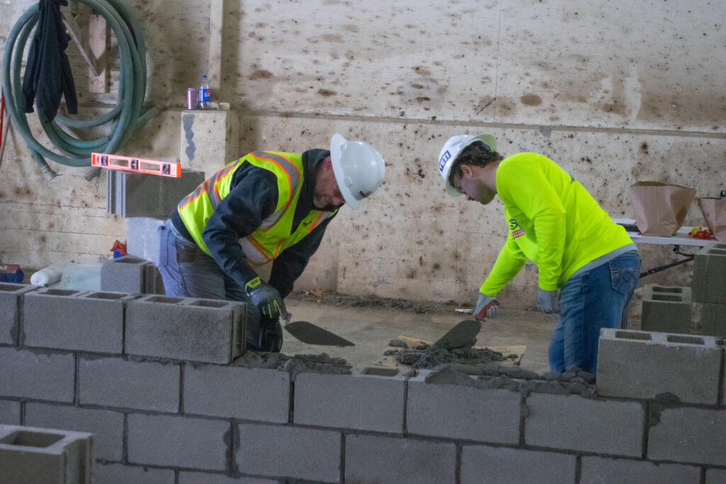 Two construction workers in safety gear and hard hats use trowels to lay bricks and mortar on a wall inside a building. A hose and soda can are visible in the background.