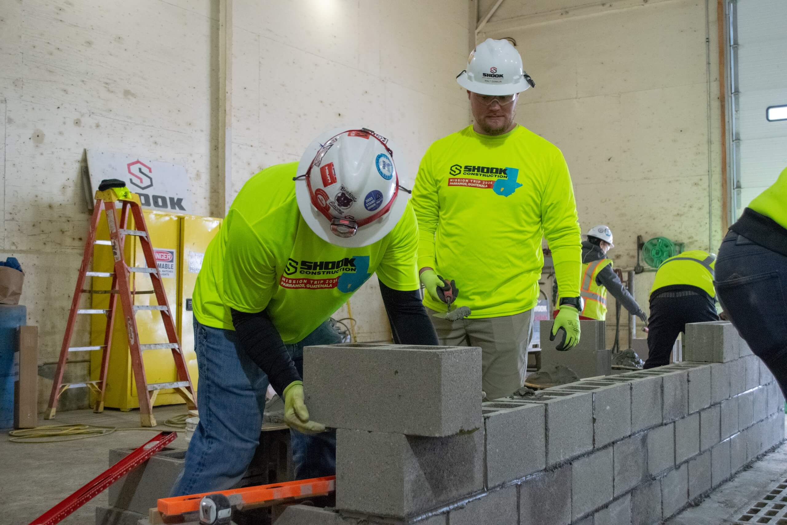 Two construction workers in bright yellow shirts and hard hats lay concrete blocks indoors, building a wall. Tools, ladders, and equipment are visible in the background.