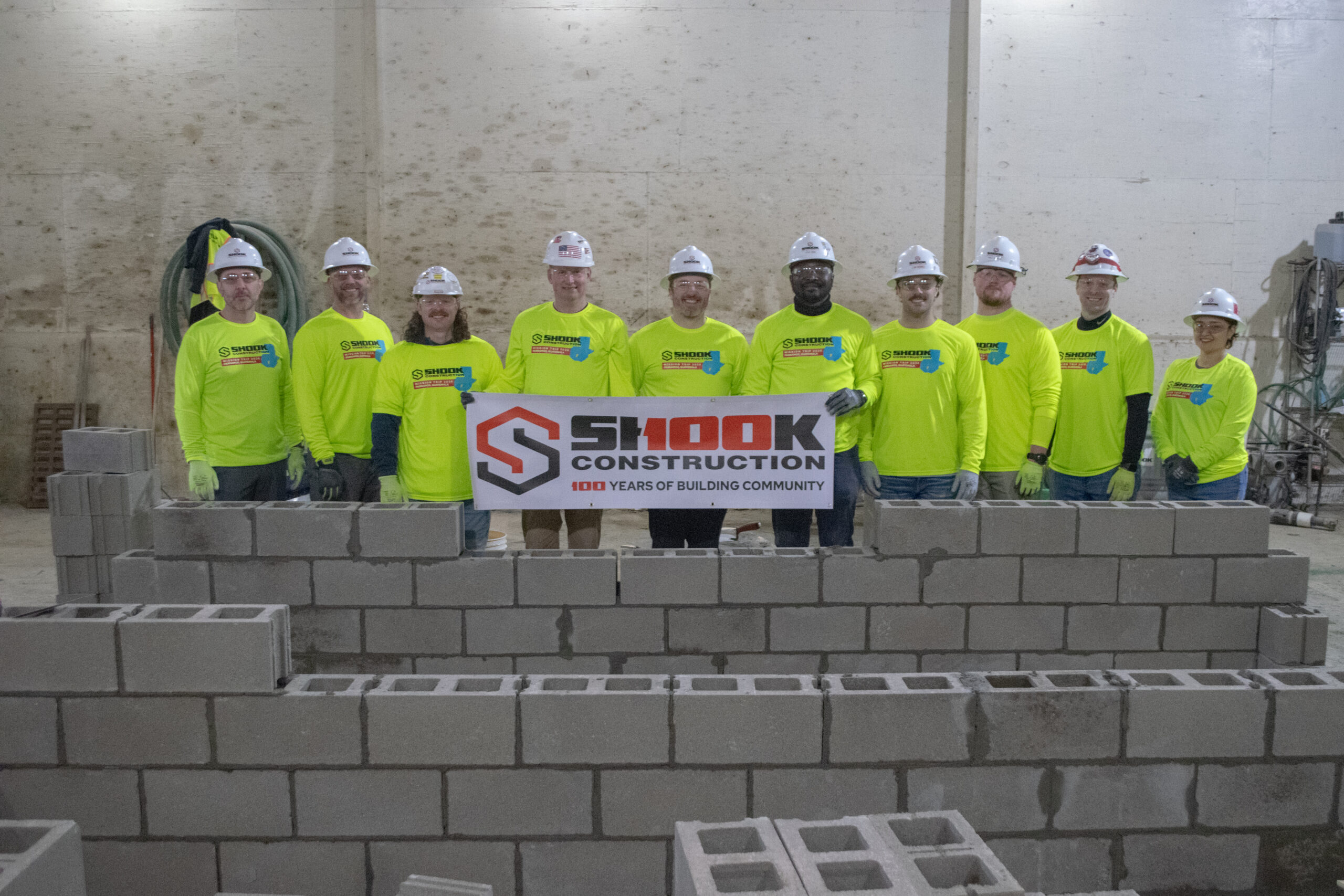 Ten people in neon yellow shirts and hard hats stand behind a low cinder block wall, holding a Shook Construction: 100 Years of Building Community sign in an industrial setting.