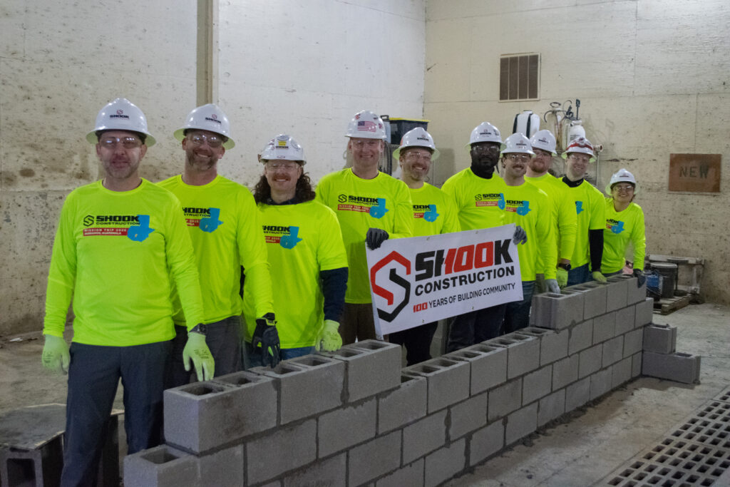 Ten people in bright yellow shirts and hard hats stand behind a row of cinderblocks, holding a Shook Construction banner in an indoor construction setting.