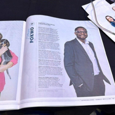 An open magazine on a table shows a woman holding a dog on the left page and a man in a suit on the right page. Another magazine is partially visible at the top right.