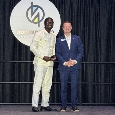 Two men stand on a stage in front of a logo backdrop. One, dressed in white, holds an award. The other wears a blue suit. Both are smiling and looking at the camera.