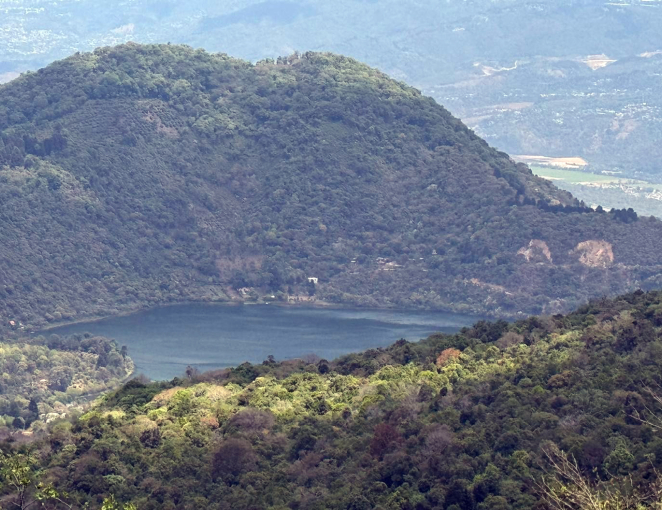 A dense green forest surrounds a small, serene lake at the base of a large, tree-covered hill, with distant mountains fading into the background under a hazy sky.