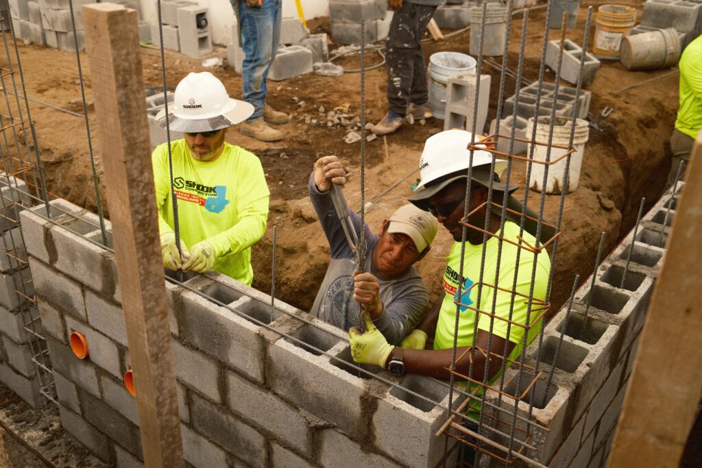 Three construction workers in hard hats and safety shirts build a cinder block wall at a construction site, handling rebar and concrete blocks in a dirt area.