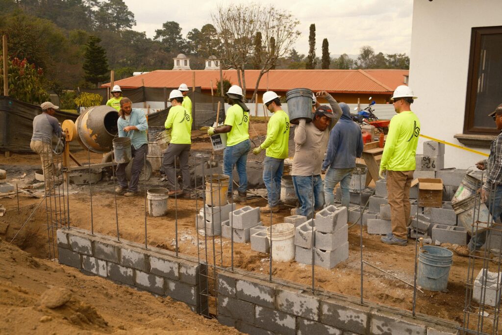 Construction workers in hard hats and safety shirts pass buckets and cinder blocks in a line at a building site, working together to construct a concrete block wall. A cement mixer and supplies are visible in the background.