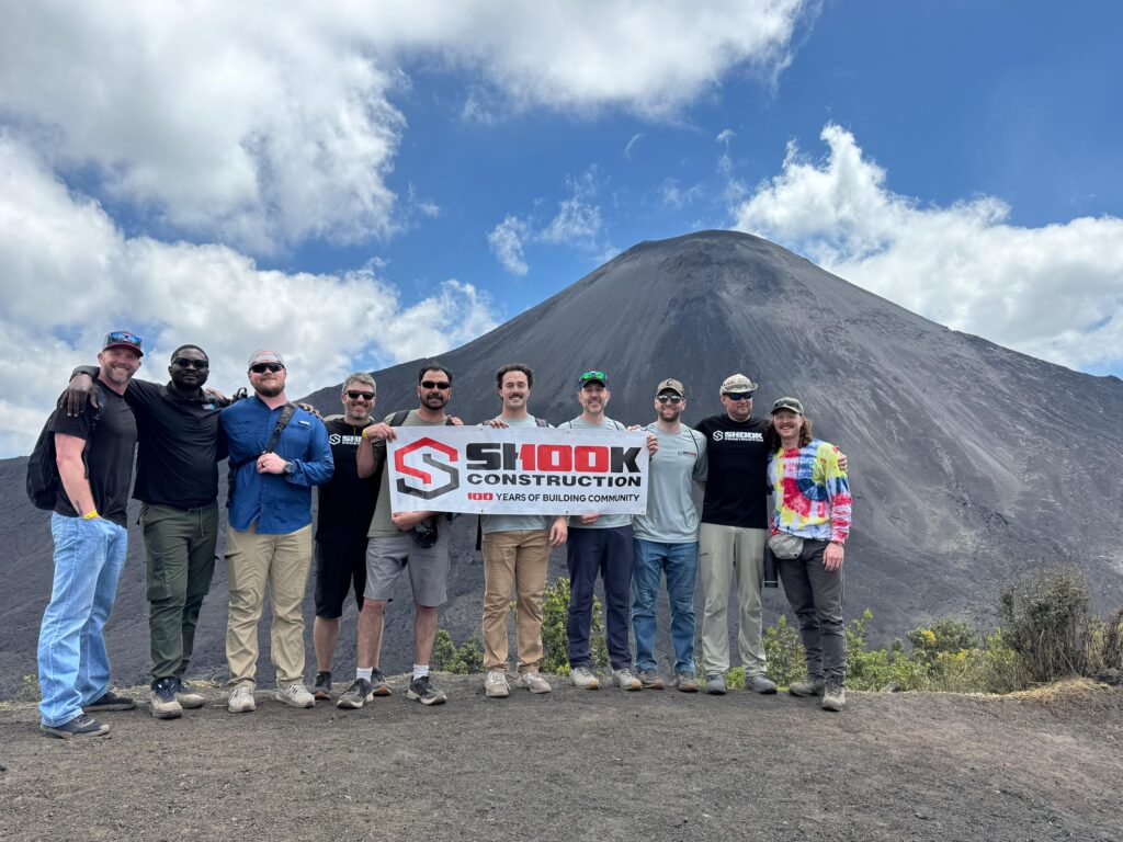 Ten people stand outdoors holding a Shook Construction banner, with a large volcano and blue sky in the background. Most are wearing casual outdoor clothing and sunglasses, smiling at the camera.