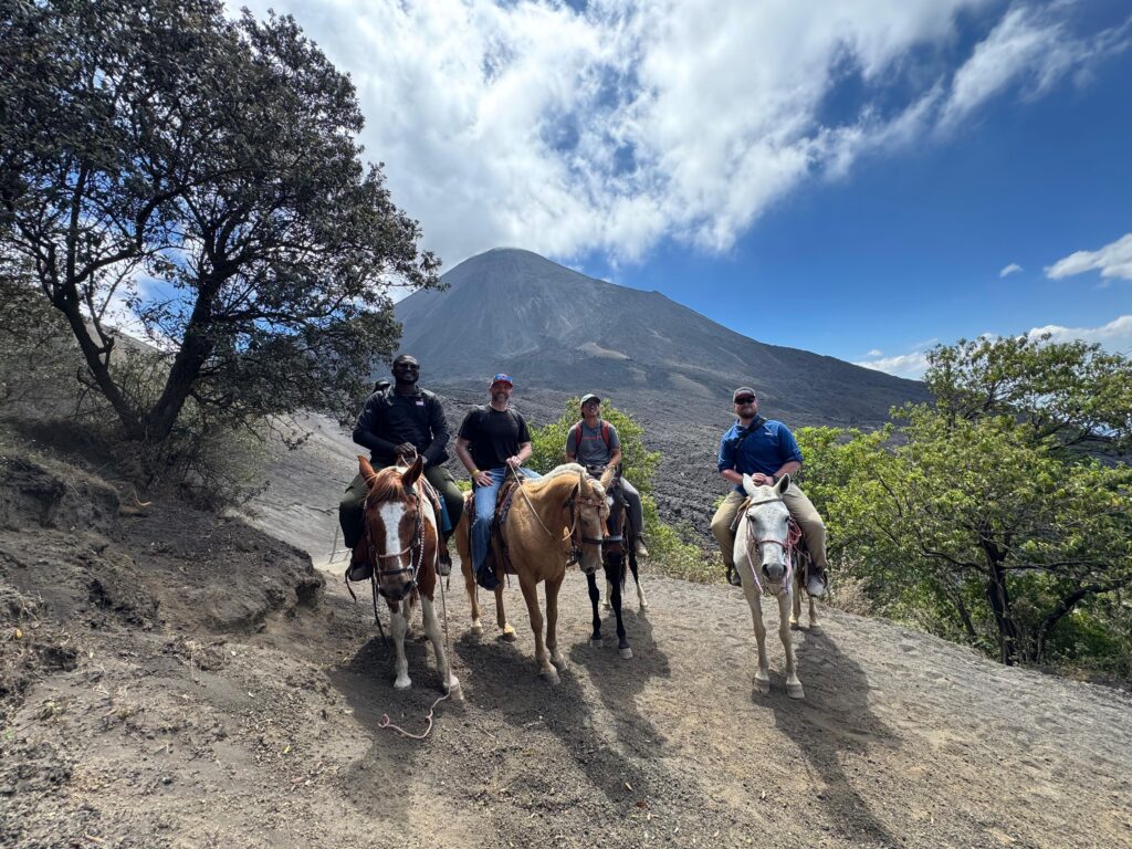 Four people on horseback pose on a dirt trail with a volcano and blue sky in the background. Trees surround the group, and sunlight highlights the rugged landscape.