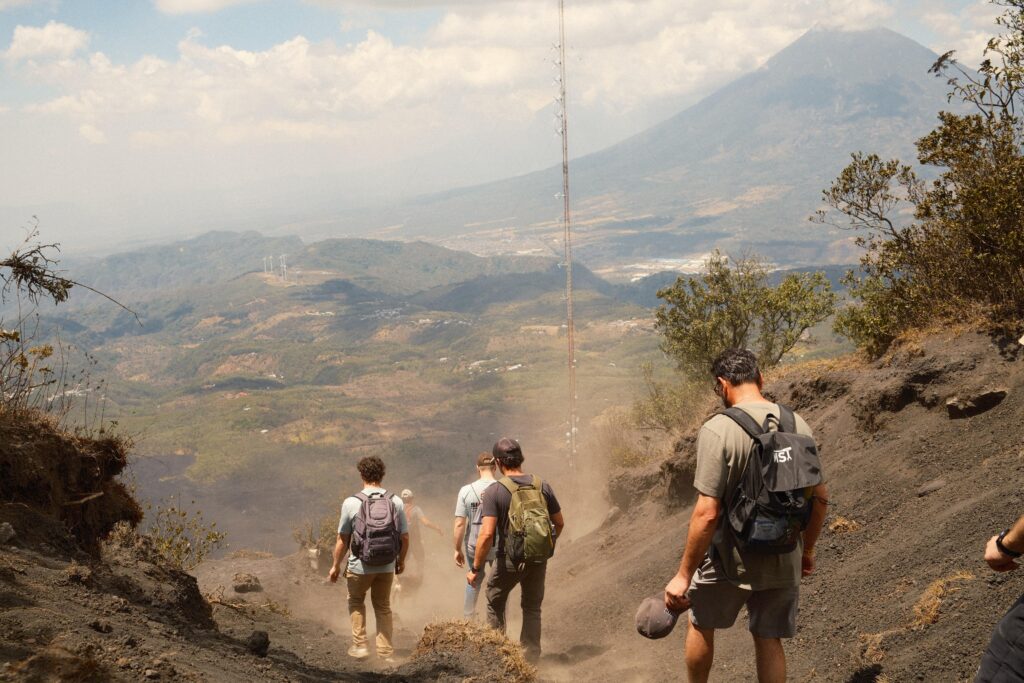 A group of hikers with backpacks walk down a dusty volcanic trail with scenic mountains and a volcano visible in the background under a partly cloudy sky.
