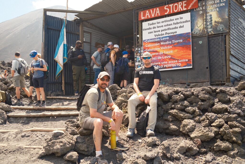 Hikers rest outside a small shop called Lava Store built from metal sheets, surrounded by volcanic rocks on a mountain trail, with more people standing near the entrance.