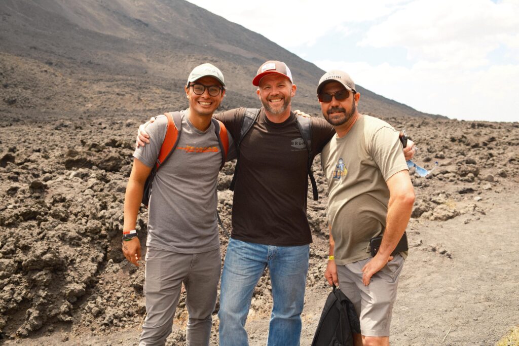 Three men stand close together, smiling, with their arms around each other in a rocky, volcanic landscape under a partly cloudy sky.