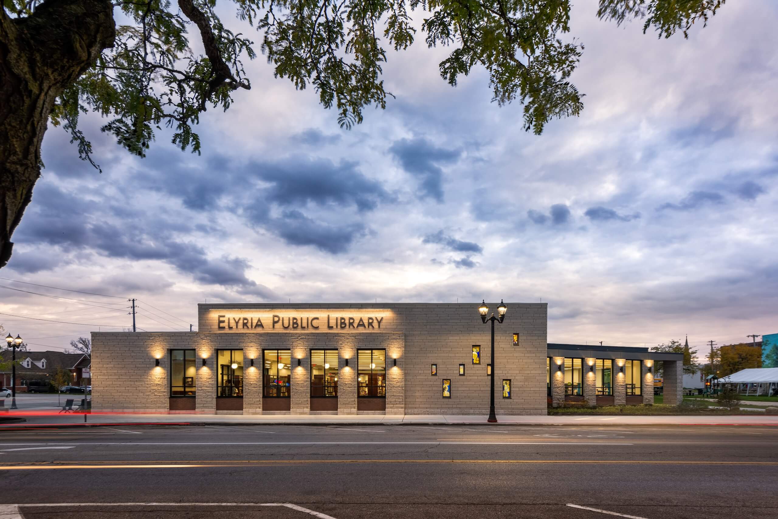 The Elyria Public Library building at dusk, with illuminated windows and sign, sits on a quiet street under a cloudy sky. Tree branches frame the top of the image.