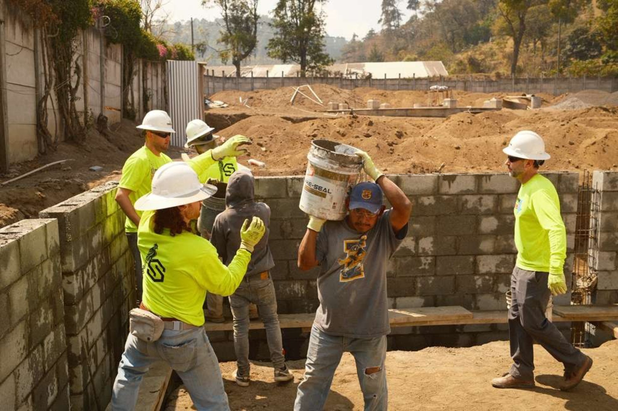 Construction workers wearing safety gear are building a cinder block wall. One worker carries a large bucket on his shoulder while others pass buckets and assist nearby. Dirt piles and equipment are visible in the background.