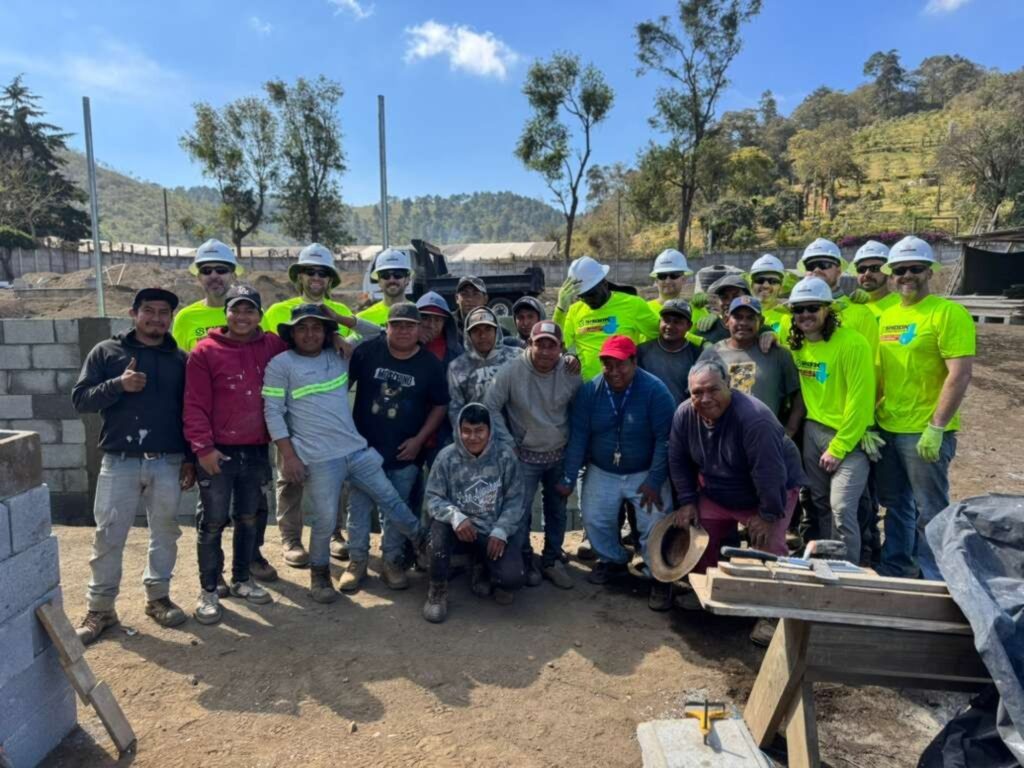 A group of construction workers pose and smile together outdoors at a building site, with trees and hills in the background. Some wear safety helmets and neon shirts.