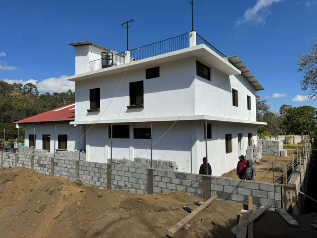 A two-story white building under construction, with a partially built concrete block wall and piles of dirt in the foreground. Several people stand near the wall, and a red-roofed structure is visible in the background.