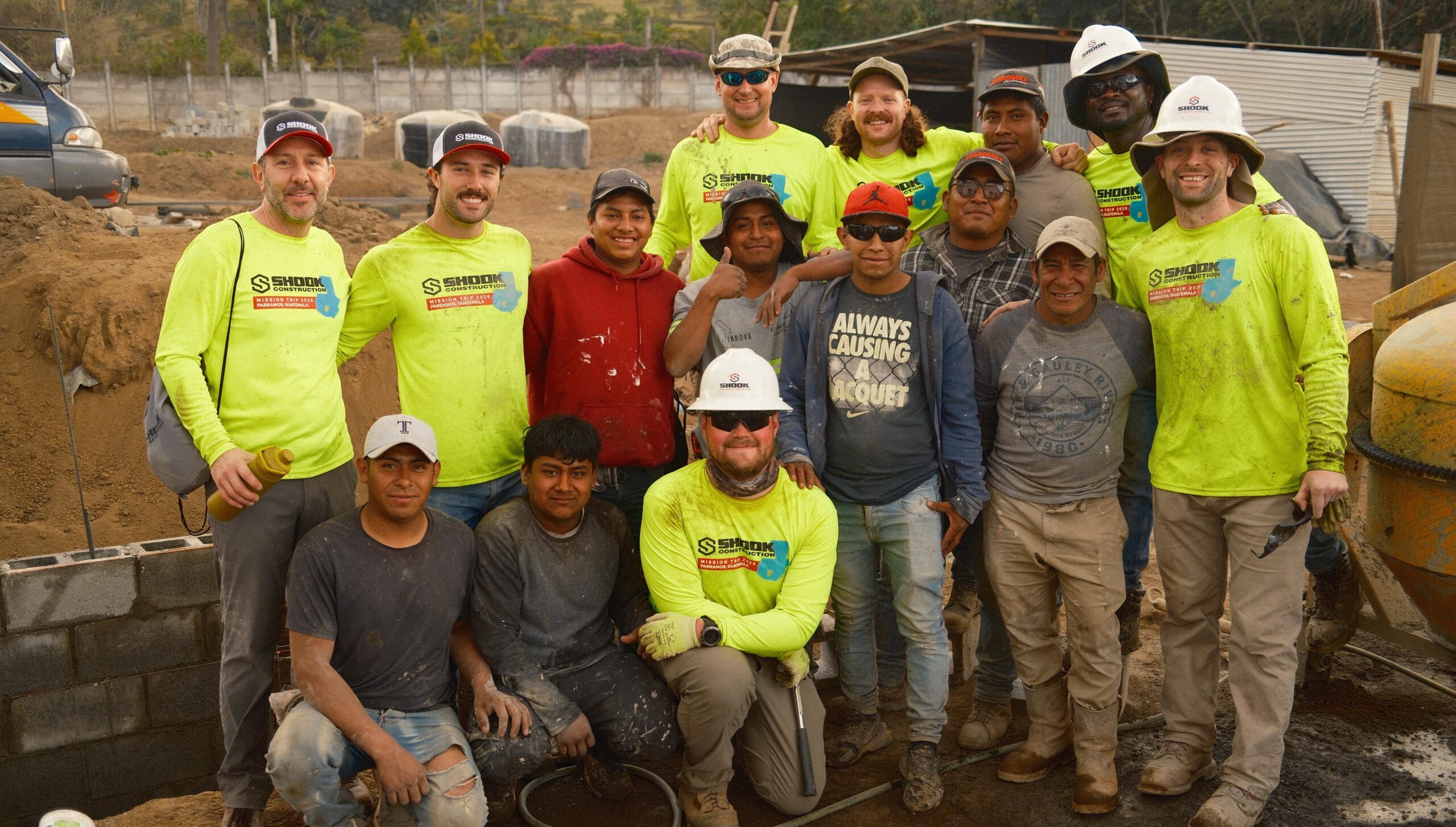 A group of construction workers, some in neon yellow shirts and hard hats, pose and smile together at a worksite with dirt, blocks, and building materials around them.