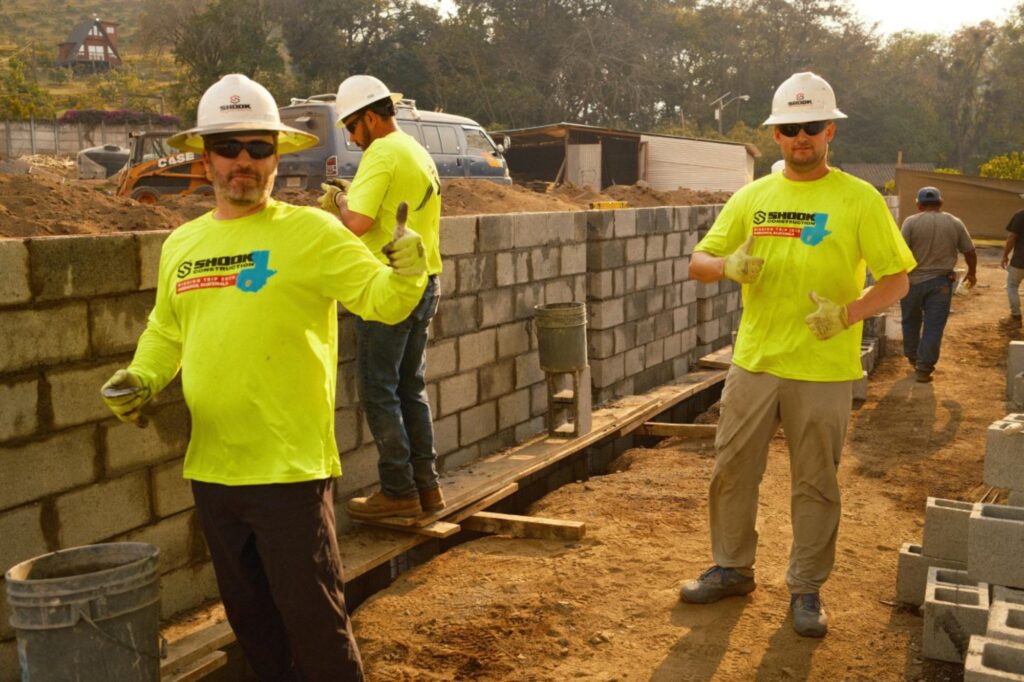 Two construction workers in yellow shirts and hard hats give thumbs up at a building site with a partially built concrete block wall and other workers in the background.