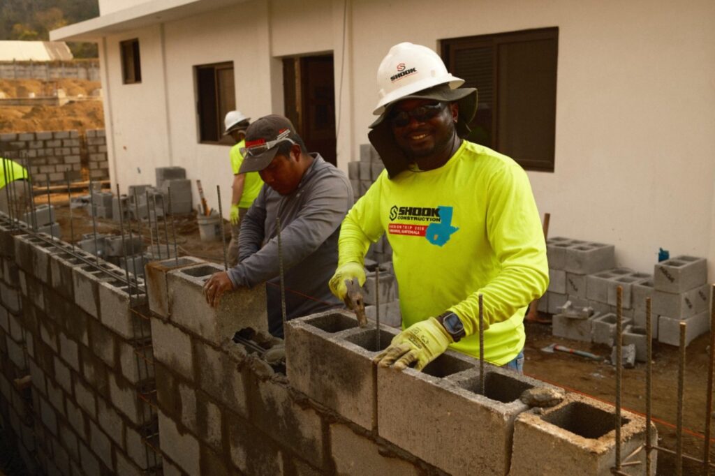 Two construction workers in safety gear build a cinder block wall at a worksite. One worker smiles at the camera while placing a block, and the other focuses on the wall. A building is visible in the background.