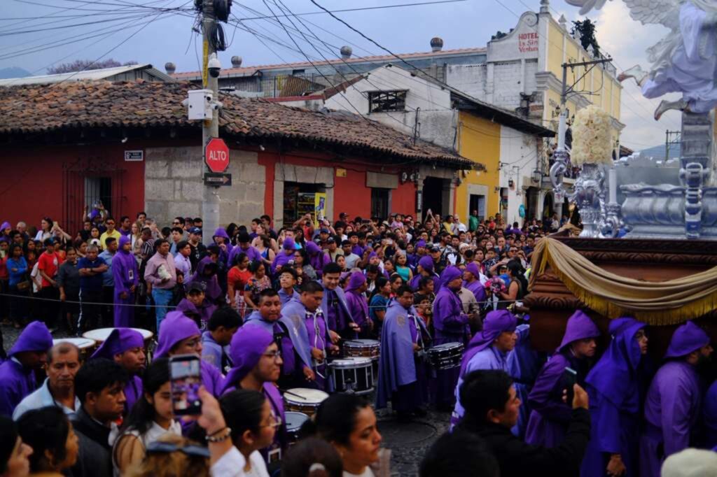 A large crowd watches a procession with people in purple robes, some playing drums, as they parade a religious float through a street lined with colonial buildings under a cloudy sky.