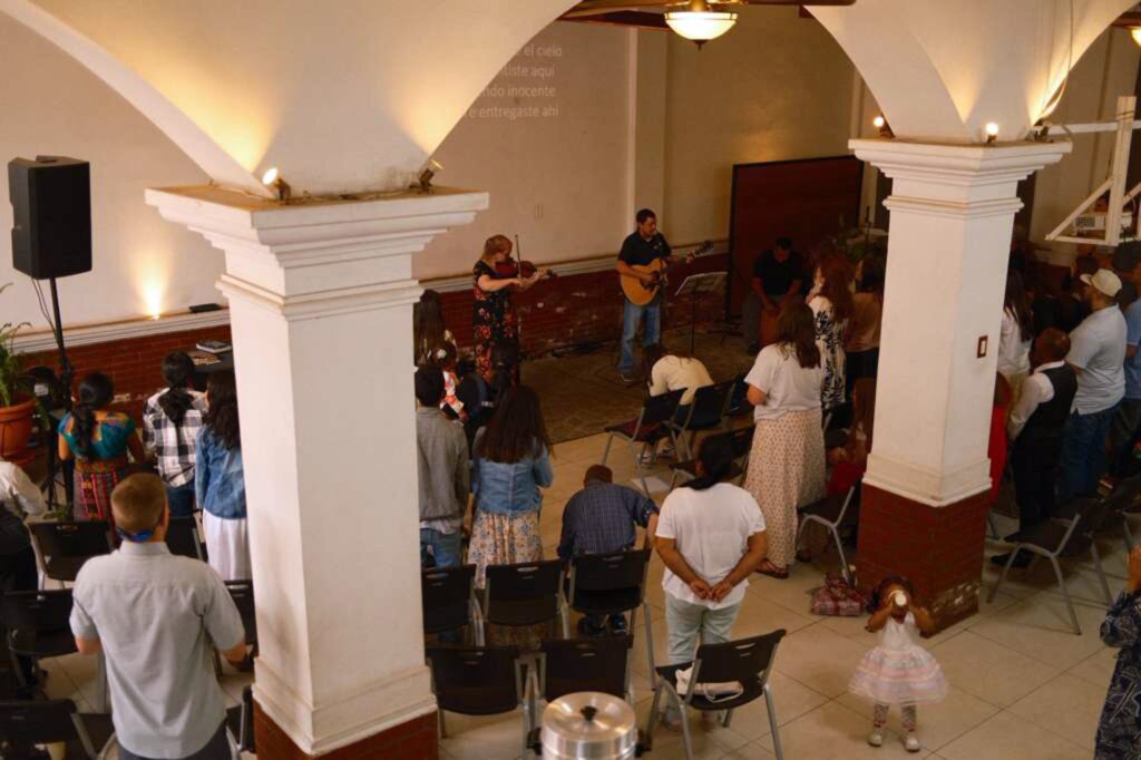 A group of people stand and face musicians playing violin and guitar at the front of a hall, engaged in a gathering or worship service. White pillars and arches are visible in the spacious, well-lit room.